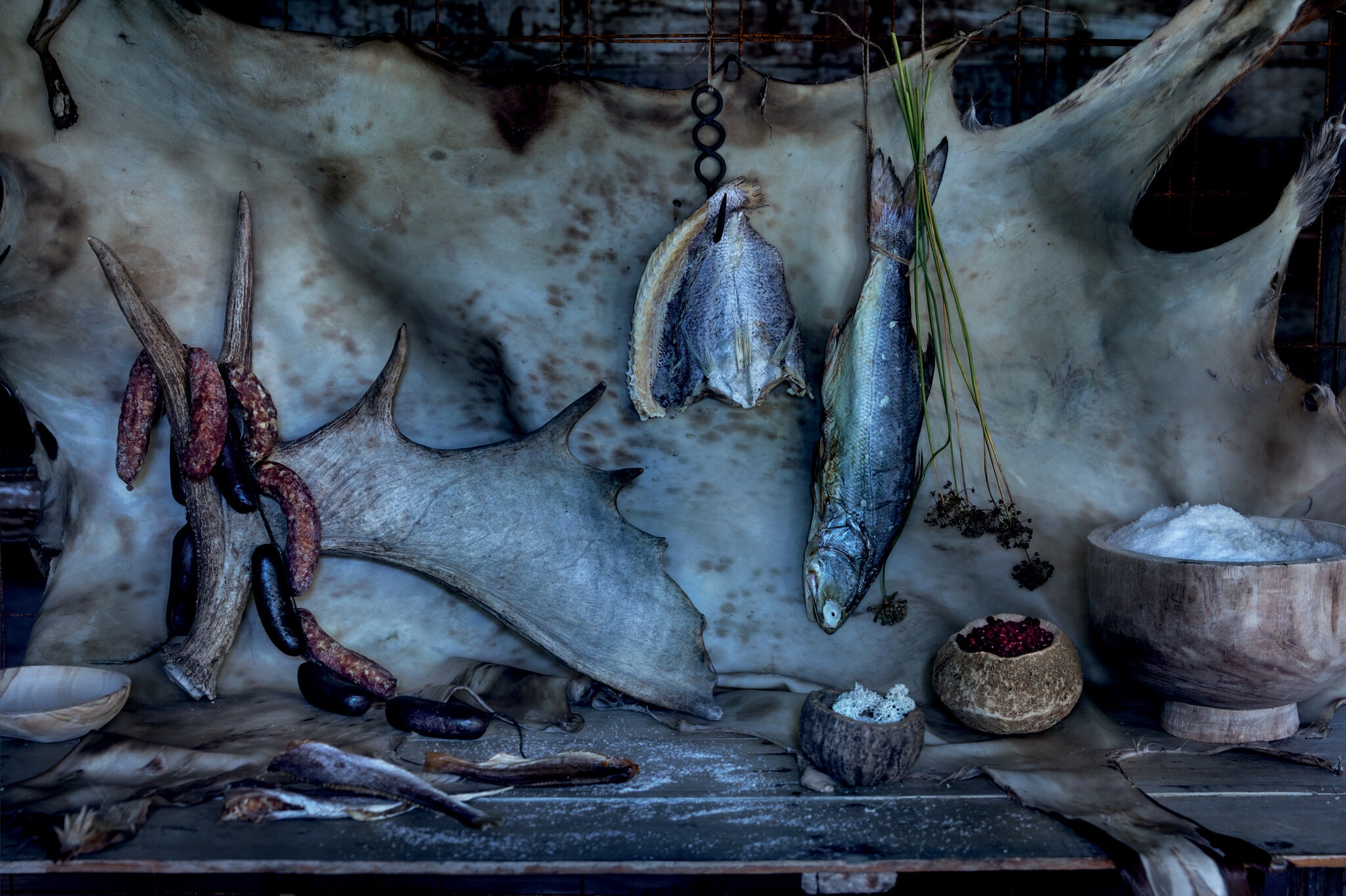 A spread of Arctic provisions including, from left, sun-dried white trout, moose antler, venison sausage, caribou blood sausage, dried Arctic flounder, dried sea bream, caribou lichen, meadow onion stems and seed heads, dried wild crowberries and geothermal Arctic sea salt against a deer hide backdrop.